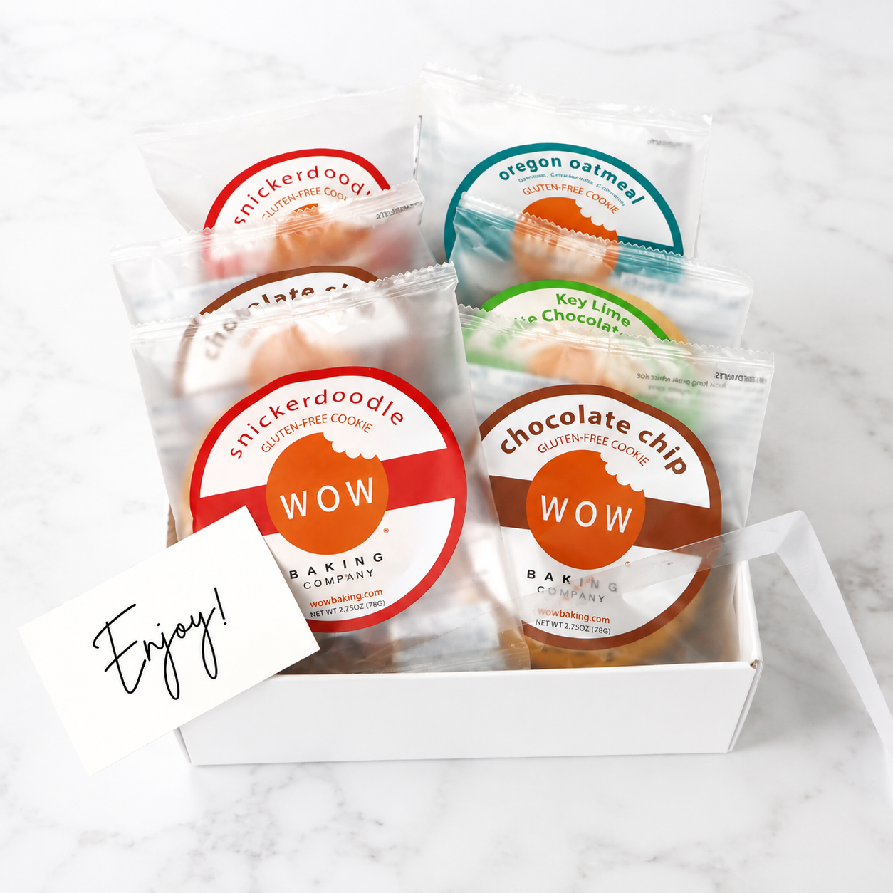 A white gift box filled with several individually wrapped WOW Baking Company gluten free cookies on a light marble surface. Visible cookie flavors include Snickerdoodle, Oregon Oatmeal, Chocolate Chip, and Key Lime Choco.
