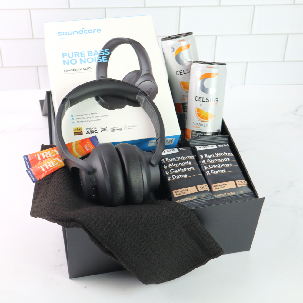 Black headphones with packaging, Celsius drinks, and protein bars in a box on a white tiled background